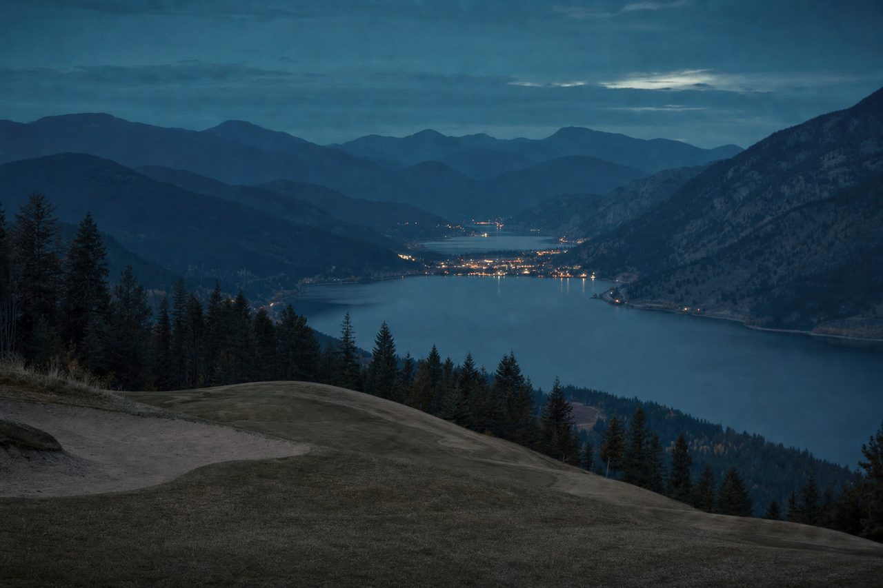 A mown golf green on a hillside at dusk, above a long dark lake with the lights of a small town along the far shore and hazy mountain ridges behind.