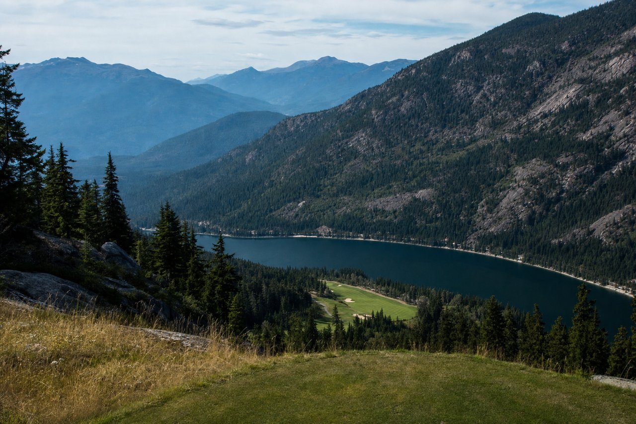 Looking down from a high tee onto a fairway far below, with a long narrow lake and hazy blue mountain ridges beyond.